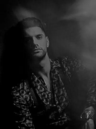Black-and-white portrait of a man with short dark hair in a patterned open shirt, seated in moody spotlight