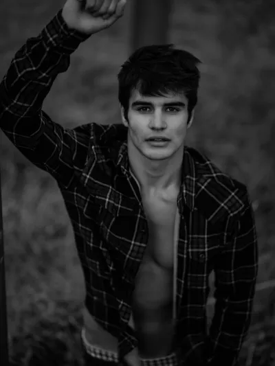 Black-and-white outdoor portrait of a young man in an open plaid shirt with one arm raised