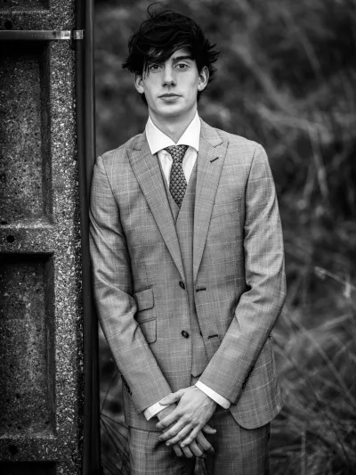 Black-and-white portrait of a young man in a checked three-piece suit leaning on a concrete pillar