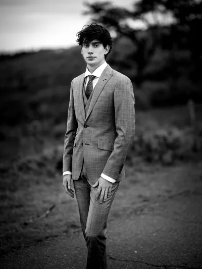 Black-and-white full-length portrait of a young man in a checked suit walking a rural path