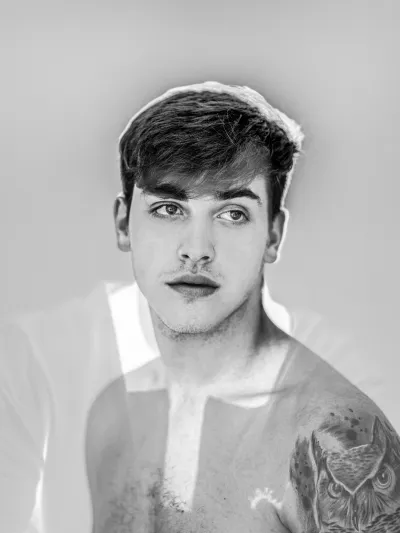 Black-and-white high-key portrait of a bare-shouldered young man with an owl tattoo on his arm