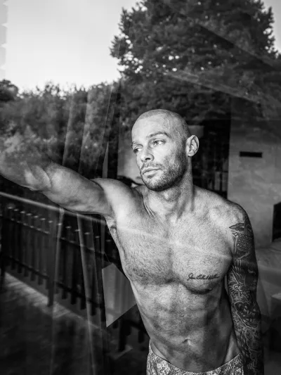 Black-and-white portrait of a muscular shirtless man with a shaved head shot through a window