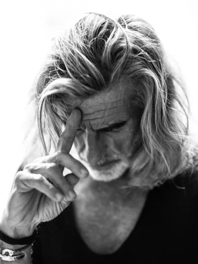 Black-and-white close portrait of an older man with long tousled hair looking down, hand to his forehead