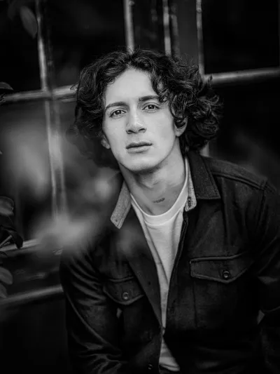 Black-and-white portrait of a young man with wavy hair in a dark overshirt, foliage bokeh
