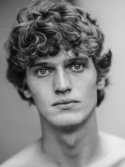 Black-and-white close-up of a young man with curly hair and light eyes, bare shoulders