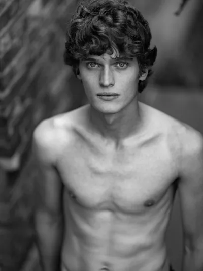 Black-and-white portrait of a slim shirtless young man with curly hair against a brick wall