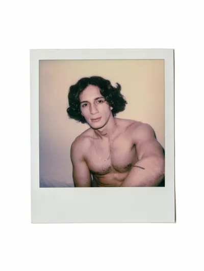 Color instant-film portrait of a shirtless young man with wavy hair and a chain necklace
