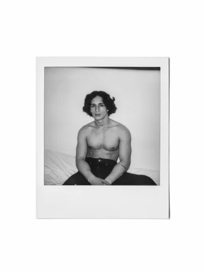 Black-and-white instant-film portrait of a shirtless young man seated on a bed