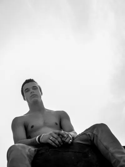 Black-and-white low-angle portrait of a shirtless young man seated against a pale sky
