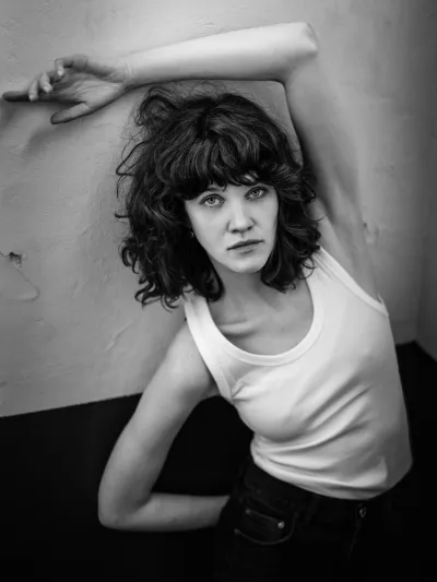 Black-and-white portrait of a woman in a white tank top with arm raised against a two-tone wall