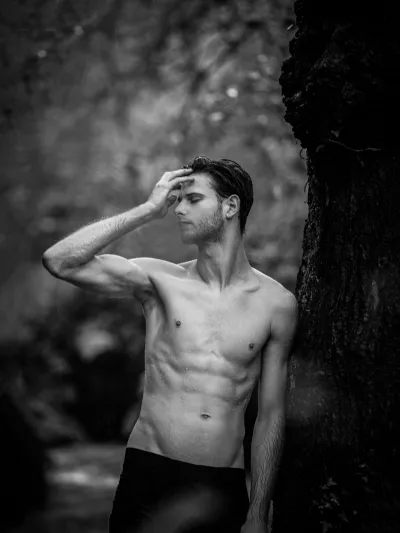 Black-and-white outdoor portrait of a shirtless man leaning on a tree, hand to his forehead