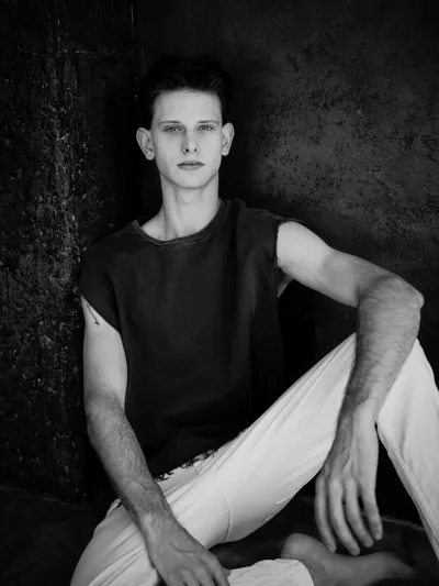 Black-and-white portrait of a young man in a sleeveless top and white trousers, seated