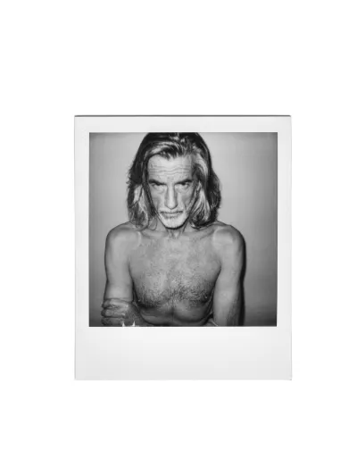 Black-and-white instant-film portrait of an older shirtless man with long grey hair, arms crossed