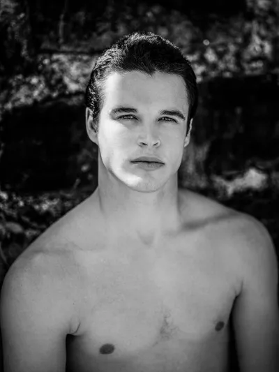 Black-and-white portrait of a shirtless young man with slicked hair against a stone wall