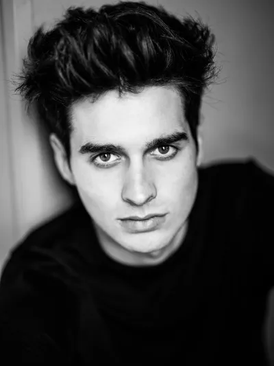 Black-and-white portrait of a young man with voluminous dark hair in a black top leaning forward