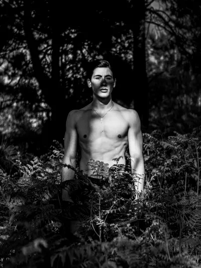 Black-and-white forest portrait of a shirtless young man standing among tall ferns