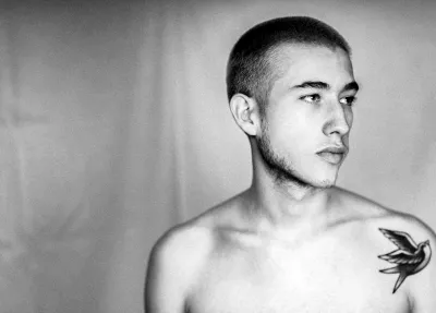 Black-and-white profile portrait of a shirtless young man with a shaved head and small chest tattoo