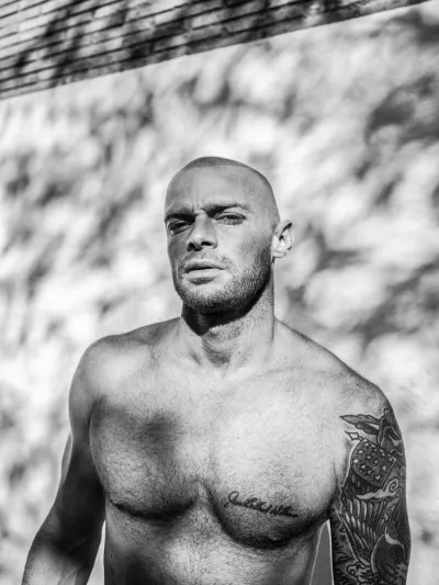 Black-and-white low-angle portrait of a shirtless bald man with chest and arm tattoos