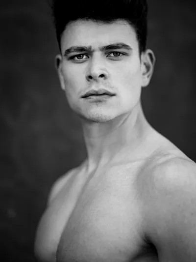 Black-and-white portrait of a shirtless young man with a dark quiff against a mottled backdrop