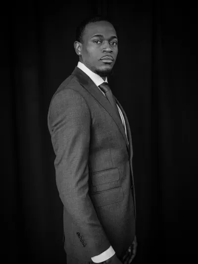 Black-and-white profile portrait of a man in a grey three-piece suit on a black backdrop