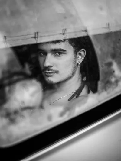 Black-and-white portrait of a young man with a moustache and lip piercing seen through a rain-flecked window