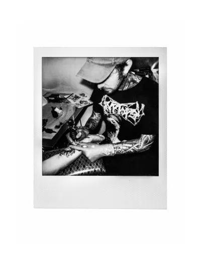 Black-and-white instant-film photo of a tattoo artist tattooing a client's forearm