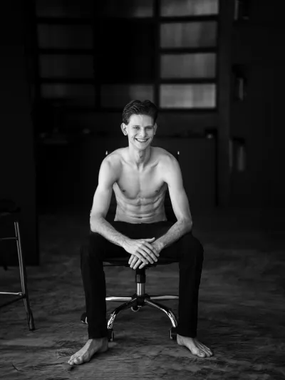 Black-and-white portrait of a smiling shirtless young man seated on a stool indoors