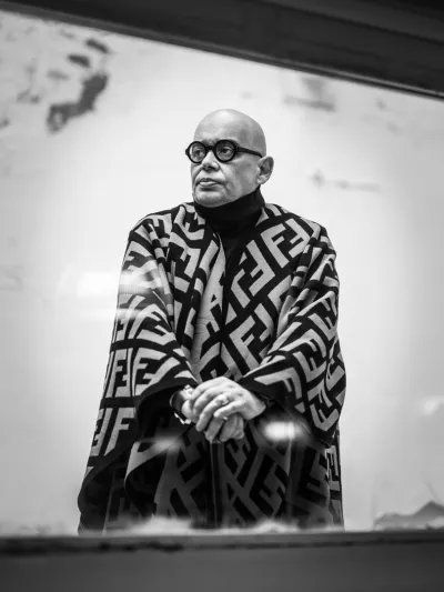 Black-and-white low-angle portrait of a bald man in round glasses and a monogram-patterned cape