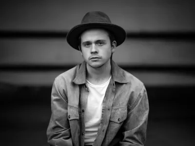 Black-and-white portrait of a young man in a wide-brimmed hat and tan jacket, seated