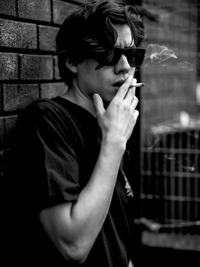 Black-and-white portrait of a young man in sunglasses smoking against a brick wall