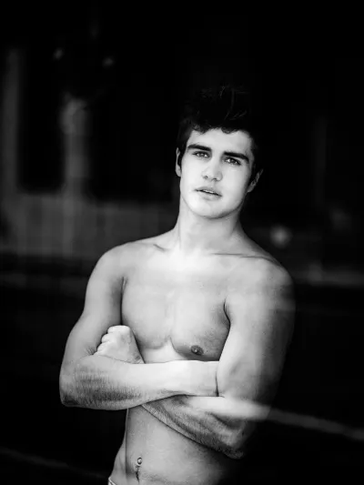 Black-and-white portrait of a shirtless young man with arms crossed and dark tousled hair